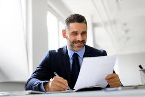 Positive middle aged businessman working with papers at workplace in office, entrepreneur man reading financial reports and taking notes, enjoying company growth and smiling photo