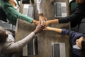 Corporate Spirit. Top View Of Diverse Coworkers Joining Hands In Stack Sitting With Laptops At Desk During Work Teambuilding Meeting In Office. Business People Stacking Arms Celebrating Achievement photo