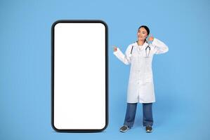 Smiling Asian Doctor Lady Gesturing Call Me While Standing Near Big Blank Smartphone, Therapist Woman Pointing At Mobile Phone With White Empty Screen, Posing On Blue Studio Background, Mockup photo