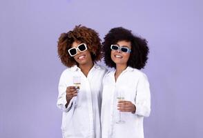 Party With Best Friend. Two Smiling Black Young Women Holding Glasses Of Sparkling Wine Standing On Purple Background, Studio Shot Of Friends Duo Toasting And Celebrating Holiday Together photo
