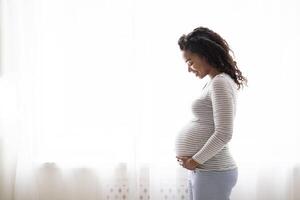 Profile Portrait Of Black Smiling Pregnant Woman Tenderly Embracing Belly While Standing Near Window At Home, Happy Expectant Mother Enjoying Her Healthy Pregnancy And Awaiting Time, Copy Space photo