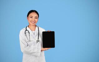 Medical App. Asian Female Doctor Showing Digital Tablet With Blank Blank Screen At Camera While Standing On Blue Background, Therapist Woman Recommending New App Or Website For Telemedicine, Mockup photo