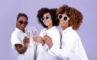 Female Bachelorette Party. Trio Of Joyful African American Ladies In Sunglasses Celebrating Holiday Clinking Glasses, Drinking Sparkling Wine Together Over Purple Studio Background. Celebration photo