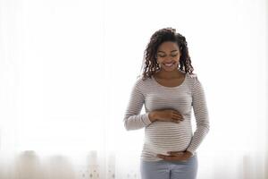 Portrait Of Beautiful Black Pregnant Woman Standing Near Window At Home, Embracing Belly. Young African American Expectant Mother Tenderly Touching Tummy And Smiling, Enjoying Pregnancy, Copy Space photo