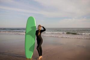 Beautiful Female Surfer Standing On The Beach And Holding Surf Board, Happy Young Woman Wearing Wetsuit Ready For Surfing, Waiting For High Waves, Looking At Camera And Smiling, Copy Space photo
