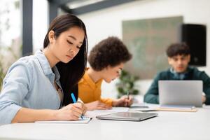 Asian lady sitting at desk and taking notes in copybook, writing essay, studying for class assignment with classmates on background. Diverse group of students learning photo