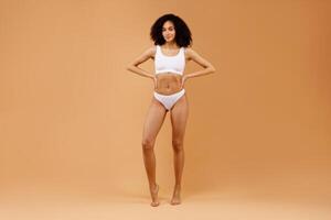 Full length shot of young beautiful latin lady posing in white underwear bra and panties over beige studio background, holding hands on her waist, demonstrating nice curves photo