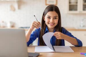 E-Learning Self Discipline. Happy School Kid Girl At Laptop Taking Notes In Textbook, Sitting At Desk Smiling To Camera At Home. Successful Schoolgirl Learning Online Excelling In Rremote Studies photo