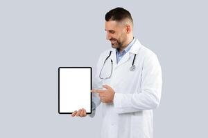 Great medical mebsite. Doctor man pointing at digital tablet with blank white screen, posing on light grey studio background. Smiling therapist showing free place for ad design, mockup photo