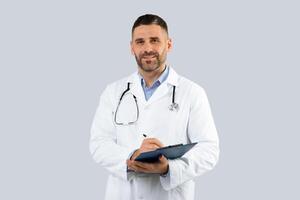 Professional doctor man in medical coat with clipboard in hands posing over light grey wall background, middle aged physician with stethoscope looking at camera photo