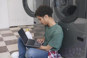 Great Laundry Service. Middle Eastern Student Guy Learning Online Via Laptop While Washing Clothes At Public Laundrette, Sitting Indoor With Computer And Surfing Web, Wearing Casual. Side View photo