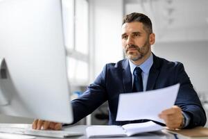 Busy middle aged male entrepreneur working on computer with documents, checking company financial reports, businessman sitting at desk in front of computer photo