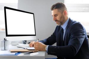 Serious middle aged businessman at table with computer with empty screen making notes, planning startup, working in office, mockup. Device for business and work, create idea photo