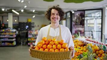 Portrait of a happy guy with curly hair as a supermarket worker who holds in his hands a large basket with a lot of tangerines and citrus fruits in the supermarket video