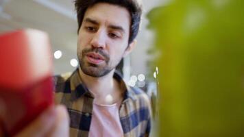 Close up a confident brunette guy with stubble in a checkered shirt approaches the counter in a supermarket and chooses the red product he needs video