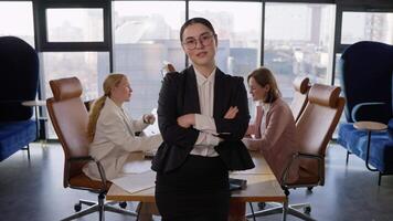 Portrait of a confident brunette businesswoman in round glasses in a black business suit who folds her arms on her chest and poses confidently in a modern office with large windows video