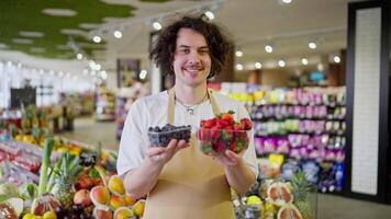 Portrait of a happy guy with curly hair a supermarket worker who holds in his hands small boxes of blueberries and strawberries in the supermarket video