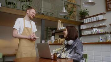 A happy guy waiter in a light brown apron takes an order from a brunette girl who is working in front of a laptop at a table in a cafe video