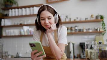 Happy and cheerful brunette waiter girl in a yellow apron looks at social networks and listens to music using white wireless headphones and a green smartphone standing at the counter in a cafe video