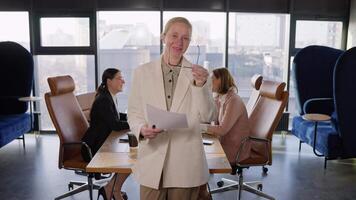 Portrait of a confident middle-aged blonde woman who takes off her glasses and poses in a white business suit near her work colleagues In the office at a table in an office with large windows video