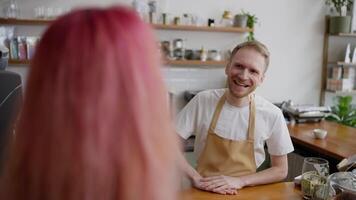 Over the shoulder of a happy blond guy in a white T-shirt and a yellow apron a barista serves a client with pink hair at the counter in a cafe video