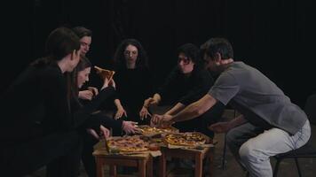 A group of happy and confident actors in black uniforms gathered around pizza having lunch and chatting during a rehearsal break on the theater stage. A group of happy actors gathered around the video