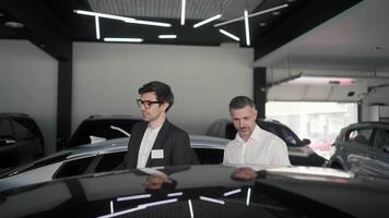 A confident brunette guy with glasses and a business suit an assistant at a car showroom tells a middle-aged man in a white shirt about modern cars at the car showroom video