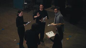 A happy and cheerful group of actors in black suits together with their stage director in a gray T-shirt and scarf are holding sheets of paper with a script in their hands and are ready to go on stage video