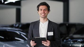 Portrait of a happy brunette man in a business suit with a tablet in the hands of a manager in a car showroom. Confident young man with a tablet in his hands posing during his work in a car dealership video