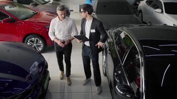 Top view of a confident brunette male assistant in a business suit talking to a middle-aged man in a white shirt about modern car models and talking about them in a car showroom video