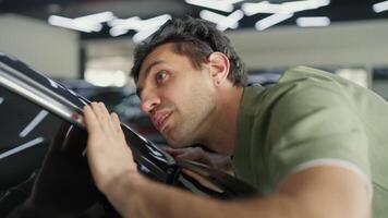 Close up a brunette guy in a green T-shirt confidently examines a modern car and wipes dust from its rear view mirror and carefully inspects the car in a car showroom video