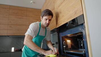 A confident brunette cleaner guy with stubble in a white T-shirt and a blue apron washes a window in an oven using a yellow rag and detergent while calling a cleaning company to the house video