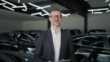 Portrait of a confident middle-aged male manager in glasses in a business suit with a tablet in his hands standing near modern black cars in a car showroom video