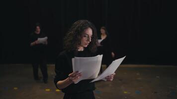 une sur de soi fille avec marron cheveux et rond des lunettes dans une noir uniforme costume lit sa mots et le terrain de le jouer de feuilles de papier pendant sa préparation et répétition sur étape dans le théâtre video