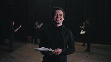 Portrait of a happy young male actor in a black suit with papers in his hands against of actors who are rehearsing for a performance on stage in a theater with black curtains video