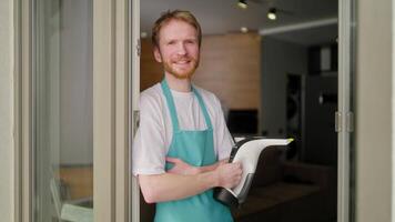 Portrait of a confident male blond cleaner with a beard in a white T-shirt and a blue apron who holds a vacuum cleaner in his hands for cleaning windows in a modern apartment video