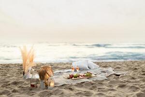 Beach picnic. Blanket with glasses of wine, snacks and fruits on the wooden board, candles on sand, beautiful view on ocean, free space. Summer evening date photo