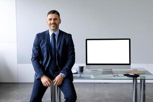Happy middle aged businessman in formal suit sitting at table with blank computer monitor, offering mockup for your design. Business ad and website concept photo