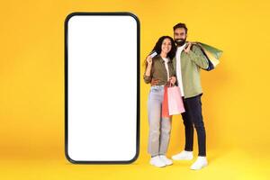 Sales Ad. Happy Arabic Buyers Couple With Shopping Bags And Credit Card Embracing Near Huge Smartphone With Empty Screen, Smiling At Camera Posing Over Yellow Studio Background. Collage photo