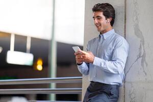 Positive handsome young man in fromal outfit employee standing at parking place next to office building, using smartphone and wireless earphones outdoors, watching content, copy space photo