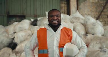 Close-up portrait of a happy and confident man with Black skin in a white protective uniform and an orange vest who is posing near a pile of recycled cellophane and plastic at a waste processing and video