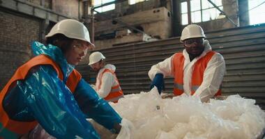 A brunette girl in a blue uniform and a white protective helmet together with her male colleagues in orange vests sorts and draws cellophane garbage at a waste processing and sorting plant video