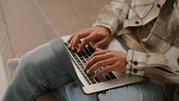 Close up of a young man with Black skin color in a beige plaid shirt and blue jeans works on a gray laptop and types on the keyboard while working remotely at home on the sofa video