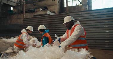 A man with Black skin in a white protective uniform in an orange vest together with his colleagues sorts and presses plastic cellophane garbage at a waste processing and sorting plant video