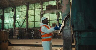 A man with Black skin in a white uniform and an orange vest with a white helmet picks up waste paper that a colleague gives him from the trunk of a truck during unloading at a waste processing plant video