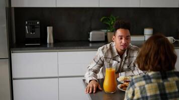 Over the shoulder a happy young guy with Black skin color a brunette in a checkered beige shirt communicates with his girlfriend with brown hair and a bob hairstyle during breakfast in a modern video
