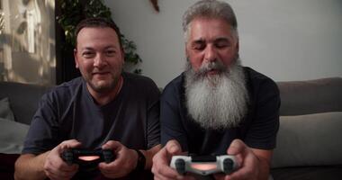 portrait of a happy man with gray hair and a lush gray beard playing games using a joystick together with his brunette boyfriend with stubble in a gray t-shirt sitting on the sofa in a modern video