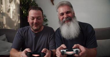 A happy brunette man with stubble in a gray T-shirt plays games using a joystick with his boyfriend. An elderly man with gray hair and a lush beard he wins and rejoices. Sitting on the sofa in a video