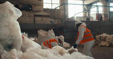 A man with Black skin in a white protective uniform and an orange vest together with his colleague, a man with a beard stacks piles of recycled cellophane in one pile at a waste processing and sorting video