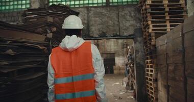 From behind, a brunette man in a white uniform and an orange vest in a White protective helmet walks along the racks with waste paper and goes out into the huge Hall of the waste recycling and sorting video